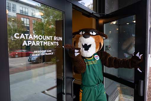 A mascot in a green jersey and brown fur coat stands in front of a glass door with the words "Catamount Run Apartments" written on it.