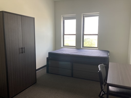 To left is a large two-door armoire. To right is a full extra long bed with exact dimensions of 54 by 80. There are two under bed drawers. To right is a desk with a desk chair.