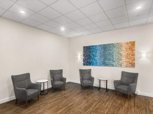 Four grey chairs are arranged in a row in a room with a large painting on the wall.