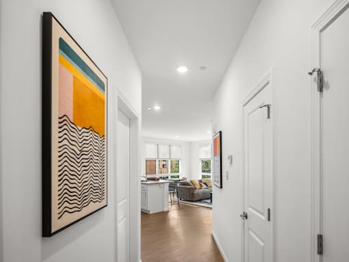 A hallway with a large painting on the wall and a view of a living room through the door.