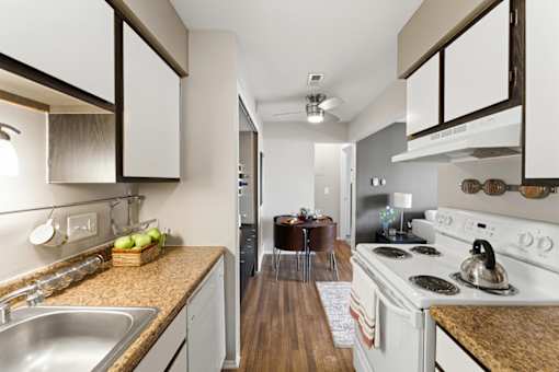 A kitchen with a white stove top oven and a sink with a bowl of fruit on the counter.