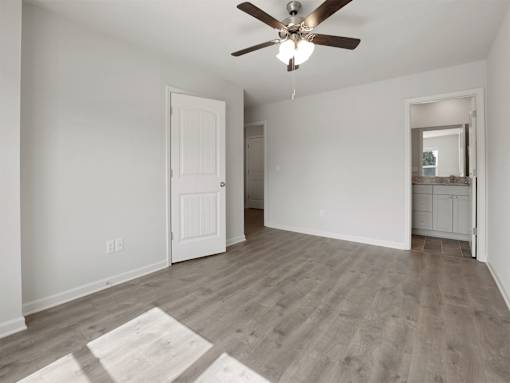 A room with a ceiling fan and light fixture, wooden flooring, and white walls with a door and a mirror.