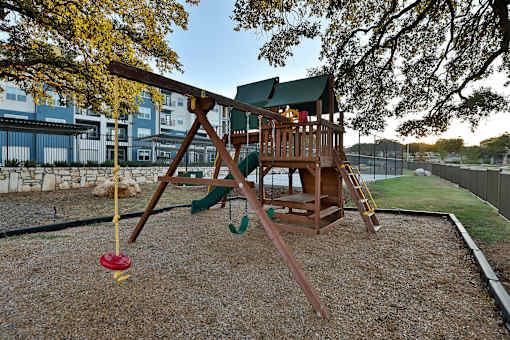 A wooden swing set with a red swing sits in a gravel backyard.