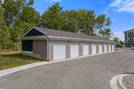 A two-story garage with a black roof and white doors is surrounded by a gravel driveway and a grassy area with trees.
