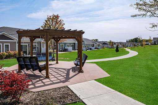 A gazebo with a bench and a trash can is in the foreground of a grassy area with houses in the background.