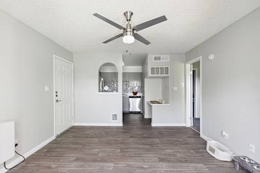 A room with a ceiling fan and a kitchen area in the background.
