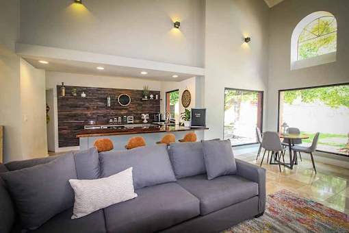 A grey couch with a white pillow is in the middle of a room with a bar area in the background.