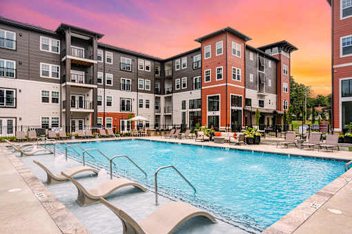 A large swimming pool is surrounded by lounge chairs in front of apartment buildings.