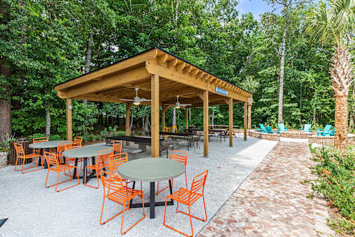 A wooden pavilion with orange chairs and a green table is surrounded by trees.