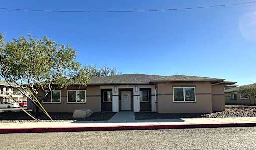 A small house with a brown roof and a white door is surrounded by a gravel driveway.