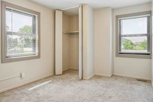 A room with a carpeted floor, two windows with blinds, and a closet with a shelf.
