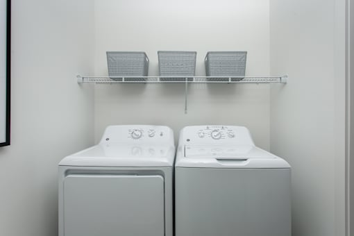 Two white washing machines are on a shelf in a laundry room.