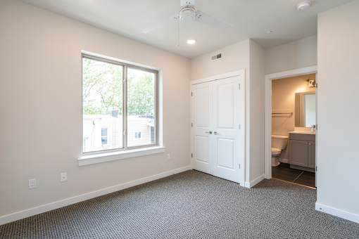 A room with a carpeted floor, a window, a door, and a bathroom visible through the doorway.