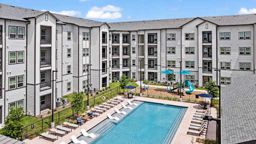 A large swimming pool surrounded by lounge chairs and umbrellas in front of a multi-story apartment building.