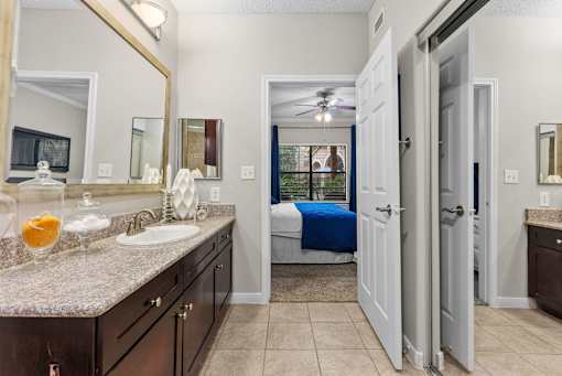 A bathroom with a sink, mirror, and a bed in the background.