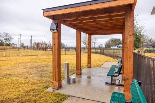 A wooden shelter with a solar panel on top is situated in a grassy area with benches.