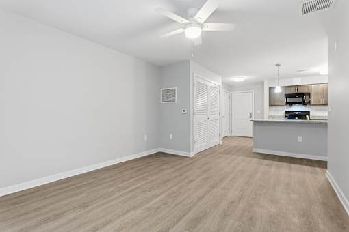 A spacious room with a ceiling fan and light, hardwood floors, and a kitchenette in the background.