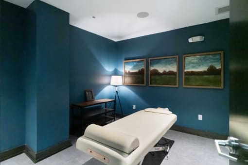 2-Bedroom Apartments in Germantown, Nashville, TN - Peyton Stakes - Massage Room with Blue Walls, White Massage Bed, Side Table with Shelves, Standing Lamp, and Wall Art.
