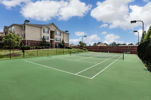 A tennis court is surrounded by a black fence and a building is in the background.