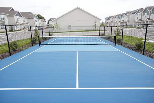 3-Bedroom Townhomes in Raymore, MO - Ascend - Fenced Pickleball Court With Blue Playing Surface, Regulation Net, And Townhome Buildings In The Background.