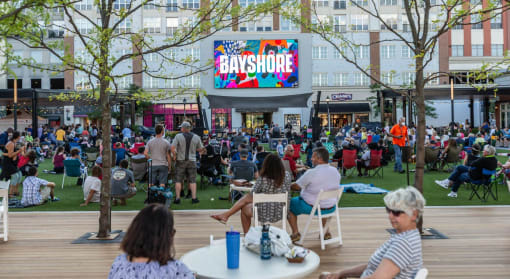 a crowd of people sit on a grassy area in front of a building with a large at The Bayshore Place Apartments in Glendale, WI