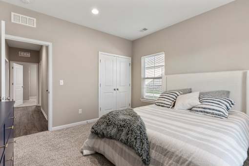 Senior Housing in Belton, MO - Traditions Villas 55+ - Bedroom with Carpet Flooring, High Ceiling, Light Fixtures, Large Bed, Dresser, Window with Blinds, and Doorway.