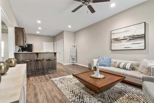 2-Bedroom Senior Townhomes in Belton, MO - Traditions Villas 55+ - Living Room with Vinyl Plank Flooring, High Ceiling, Ceiling Fan, Couch, Coffee Table, Painting, Large Rug, and a View of the Kitchen