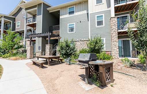 A wooden picnic table is in front of a building with a grill on it.