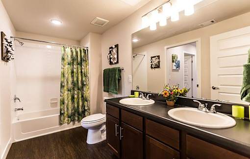 A bathroom with a white toilet, a white bathtub with a green shower curtain, a wooden vanity with two sinks, and a mirror above the vanity.