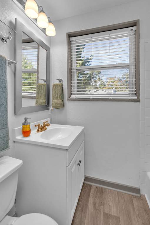 A white bathroom with a toilet, sink, mirror, and window with blinds.