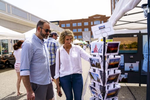a man and a woman walk past a postcard stand at a farmers market at The Bayshore Place Apartments in Glendale, WI
