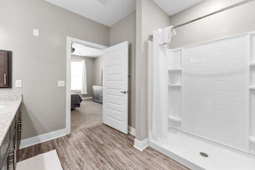 A white bathroom with a walk-in shower and a bedroom visible through the door.