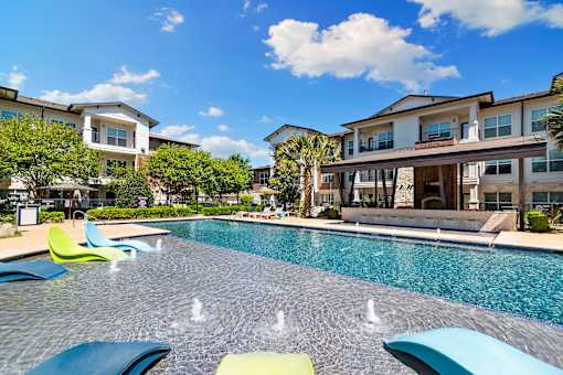 A pool with blue and green lounge chairs in front of apartment buildings.