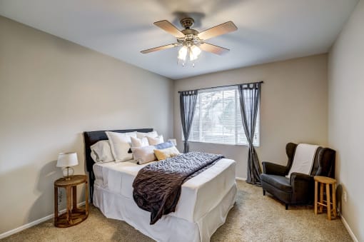 One, Two, And Three-Bedroom Apartments In Aurora, CO - Sonoma Resort At Saddle Rock - Bedroom With Bed, Chair, Nightstand, Lamp, Ceiling Fan, And Window.