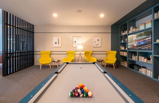 A pool table with a yellow chair and a blue bookshelf.
