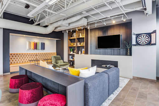 A modern living room with a grey sofa, a yellow cushion, a grey armchair, a red ottoman, a white couch, a TV, a fireplace, a dartboard, and a shelf with plants