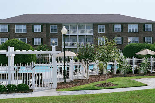 A white picket fence surrounds a pool area in front of a brick building.