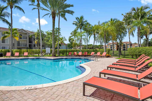 Outdoor view of large resort-style swimming pool with lounge chairs along deck and tables with chairs and umbrellas, and clubhouse in back