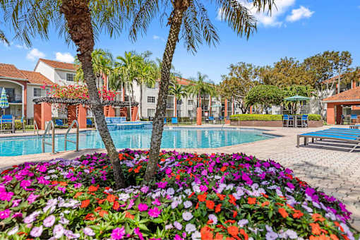 Alternate view of community swimming pool at Sunset Gardens. Hot tub is located near covered lounge area. Lounge seating is located throughout pool deck as well as picnic tables