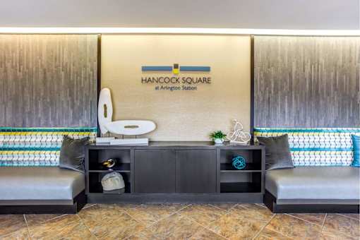 A Hancock Square at Arlington Station sign is on a wall above a shelf with a toilet paper holder and a plant.
