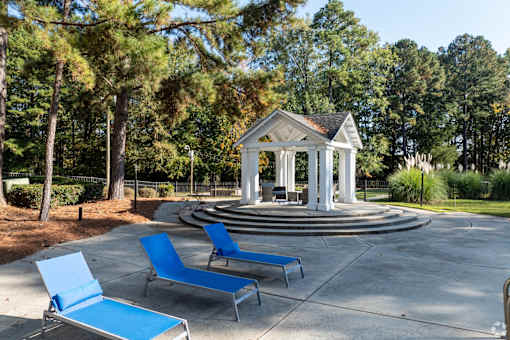 A gazebo is surrounded by trees and has two blue lounge chairs in front of it.