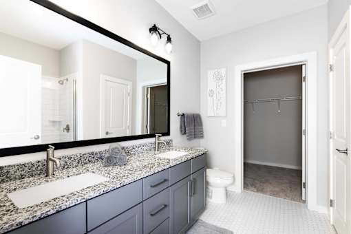 Apartments in Nashville, TN - The Sound at Pennington Bend - Bathroom with Granite Countertops, Gray Cabinets, Large Mirror, White Tile Flooring, and an Oversized Closet with Carpet Flooring.