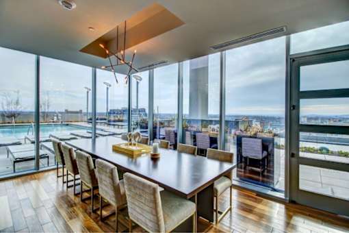 A modern dining room with a long wooden table and chairs, a chandelier, and a view of the outdoors through large windows.