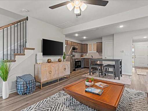 Apartments for Rent in Spring Hill, KS - Oak Woods - Living Room with Open-Concept Layout, Plank Flooring, Area Rug, Coffee Table, Ceiling Fan, Storage Cabinet, Wall-Mounted TV, and View of Kitchen Area.