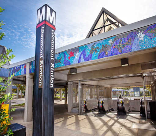 A tall pillar with the letters MM on it stands in front of a building with a mosaic on the roof.
