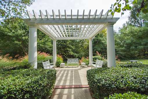 A white pergola with benches is surrounded by green bushes.