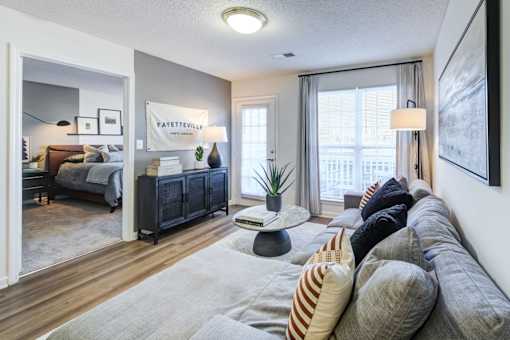 A living room with a grey sofa and a bedroom with a bed and a nightstand in the background.