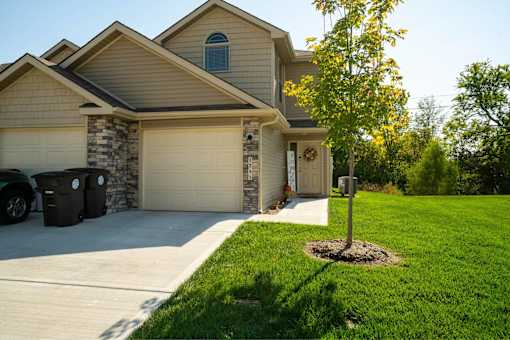 2-Bedroom Townhomes in Belton, MO - Traditions Townhomes - A Garage Attached to the House, with Driveway, Black Garbage Bins, and Lush Landscaping.