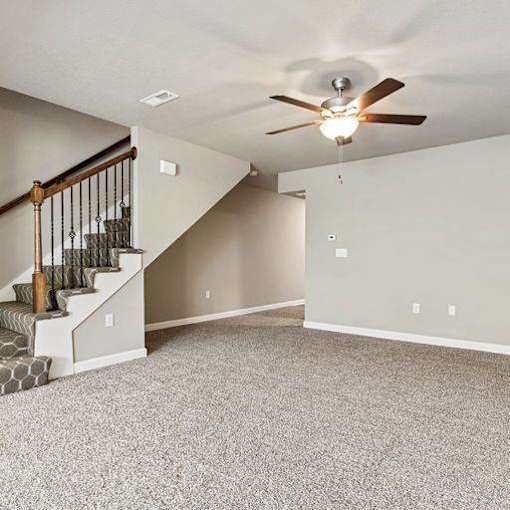 3-Bedroom Townhomes in Belton, MO - Traditions Townhomes - A Spacious Empty Room with Carpet Flooring, Ceiling Fan with Light, and a Carpeted Staircase.
