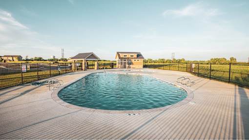 Pet-Friendly Townhomes in Belton, MO - Traditions Townhomes - An Outdoor Swimming Pool Surrounded by a Deck, Blank Fence, and Landscaping, Overlooking a Pavilion and the Clubhouse.